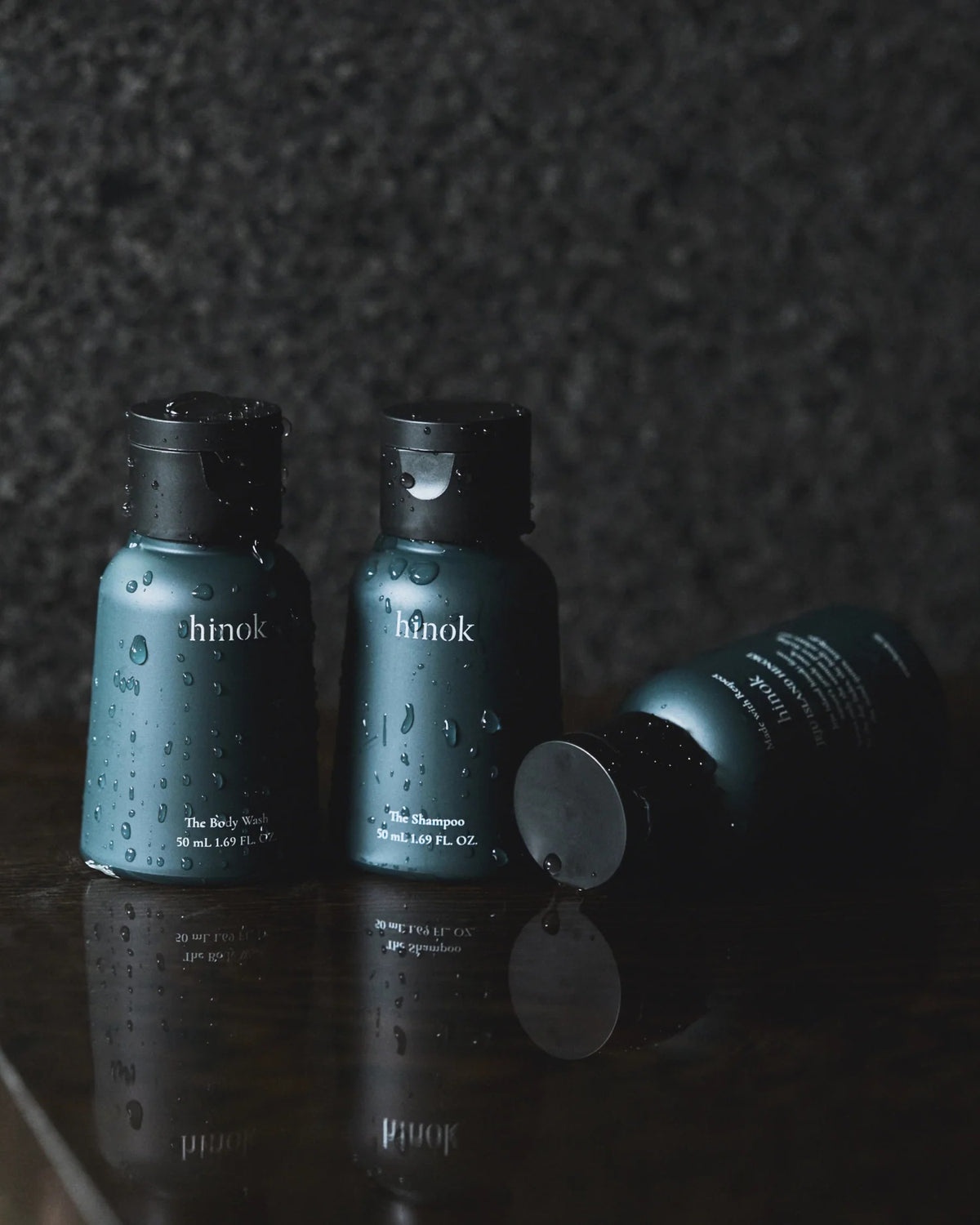 Three travel-sized bottles of different hinok hair and body formulas, covered with beads of water.
